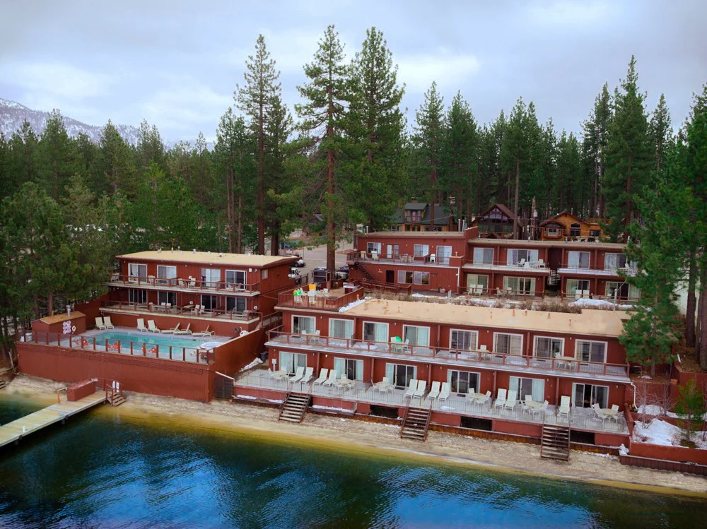 The Beachcomber Inn from the lake, showing the property's pool deck, lake-facing rooms, and pine-tree backdrop
